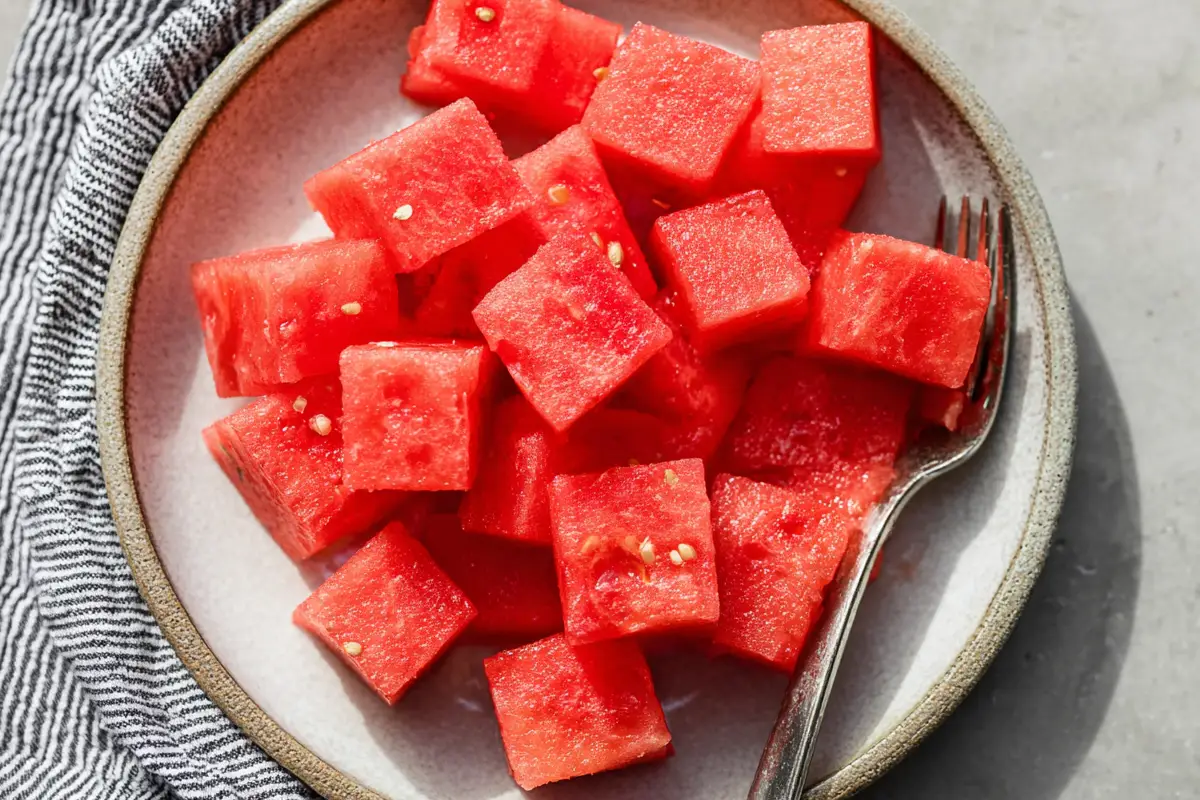 Plate of freshly cut watermelon cubes with visible seeds, served with a fork on a modern ceramic dish and a striped cloth napkin beside it